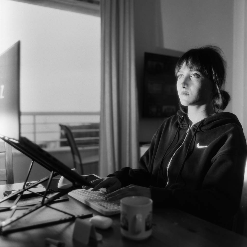 A woman works at a computer, her face lit by the glow of the monitor. She has a vacant, tired expression
