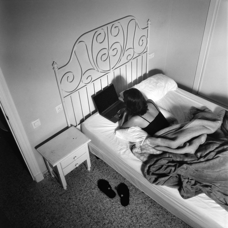 A woman works on a laptop while lying in bed. Artificial lighting suggests a late hour