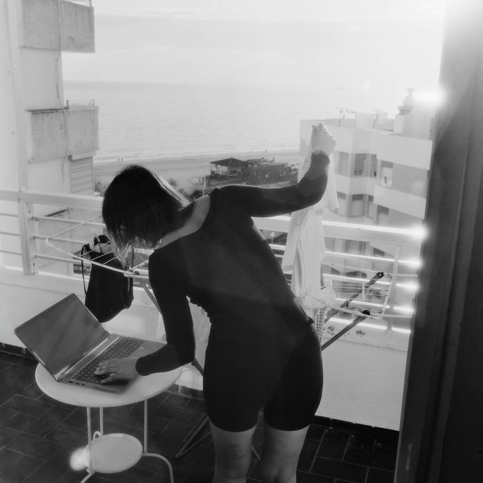 A woman hangs laundry on a balcony with one hand while typing on a laptop with the other. The sun rises in the background