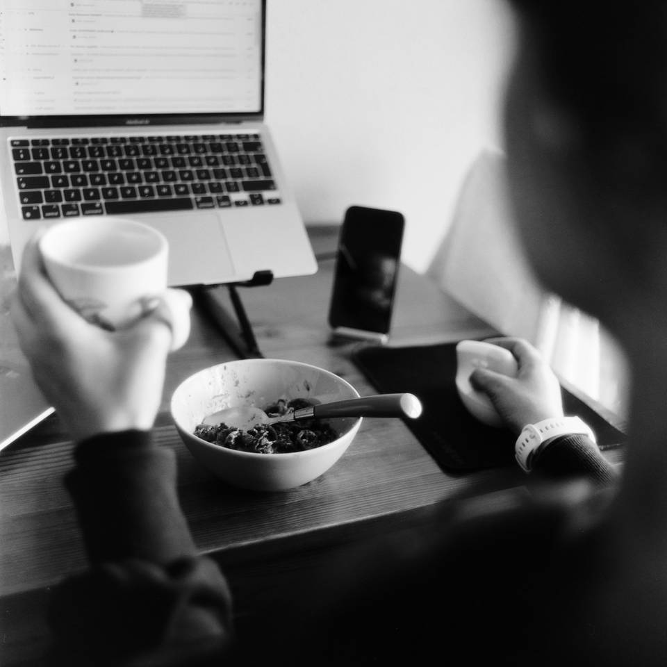 A woman sits in front of a laptop eating breakfast, holding a cup of coffee